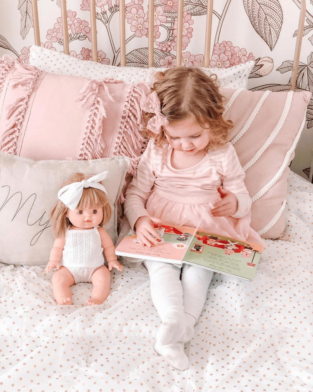 A young girl in a pink dress sits on a bed reading the Ditty Bird "Funny Songs" music book, with a doll in white beside her. The bed has pink and white pillows and floral wallpaper can be seen in the background.
