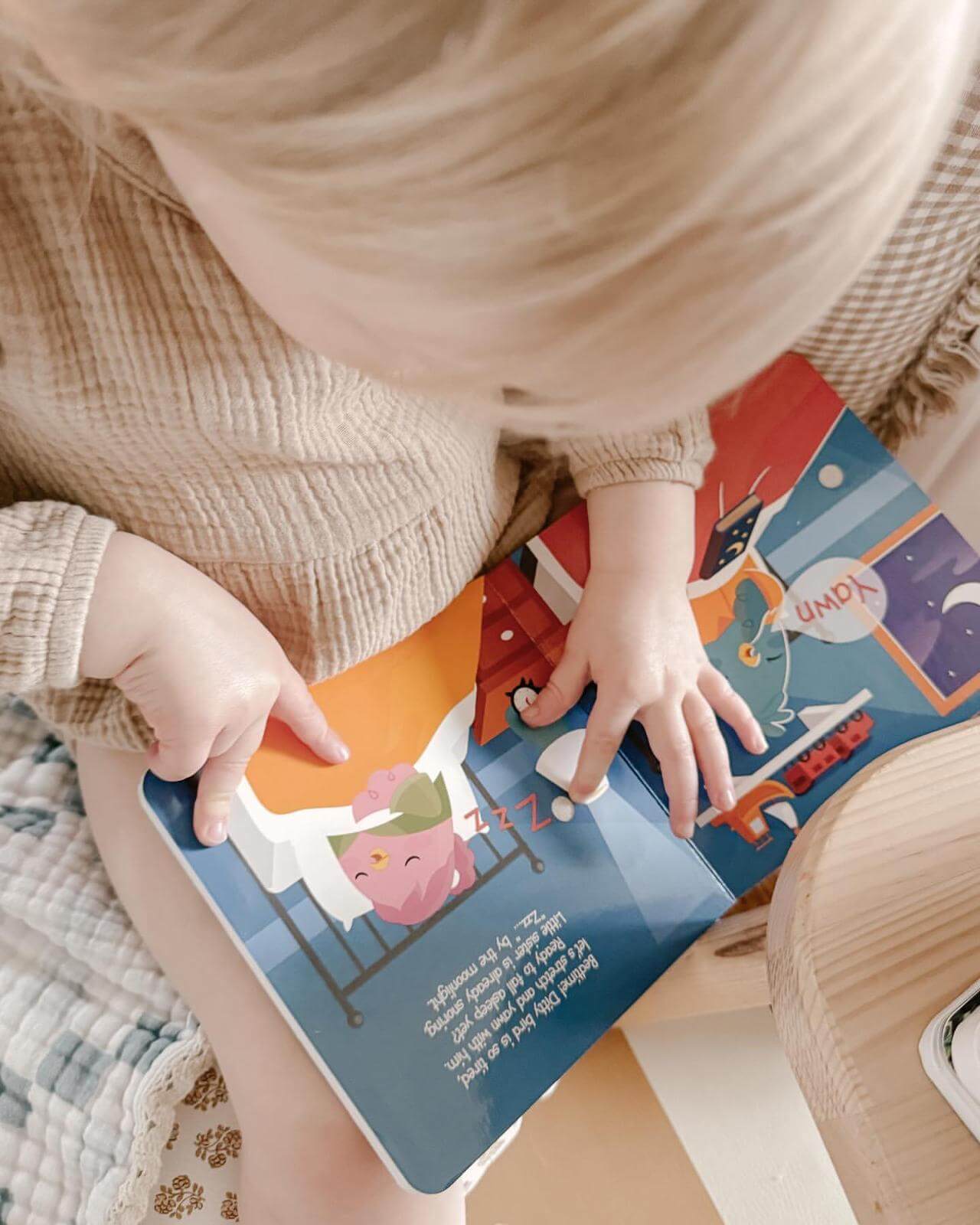 A young child in a beige textured outfit sits and uses their finger to follow along while reading Ditty Bird Potty Time Sounds | AG10, a potty training book with cartoon animals and bright backgrounds that make potty time fun and engaging.