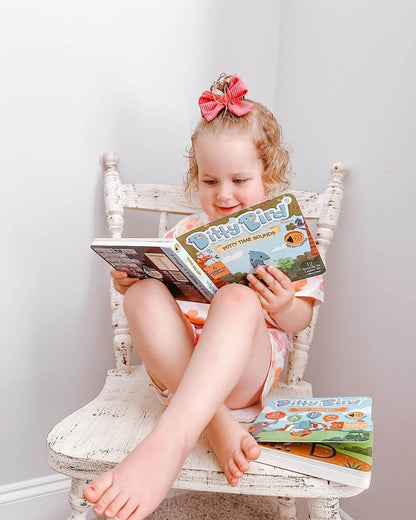 A young child with curly blonde hair and a pink bow sits barefoot on a white wooden chair, smiling as she reads the Ditty Bird Potty Time Sounds | AG10 book. Another Ditty Bird book rests on the seat beside her.