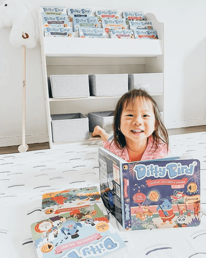 A smiling young girl lies on a bed holding Ditty Bird’s Chinese Songs Vol.2 book, with more Ditty Bird books spread in front of her and a shelf with gray bins and additional books visible in the background.