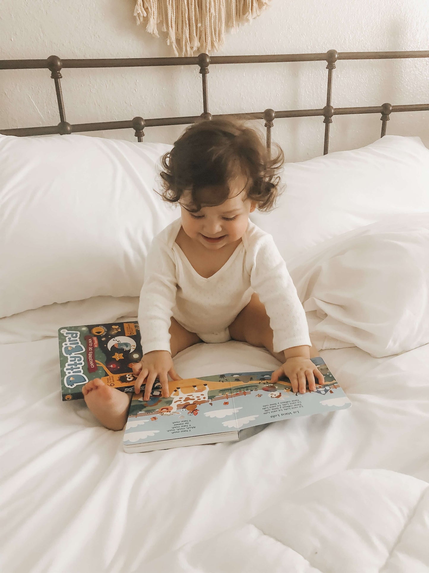 A toddler with curly hair in a white onesie smiles on a white bed while enjoying Ditty Bird's "Canciones de Cuna en Español" bilingual sound book. Another book rests nearby on the sheets.