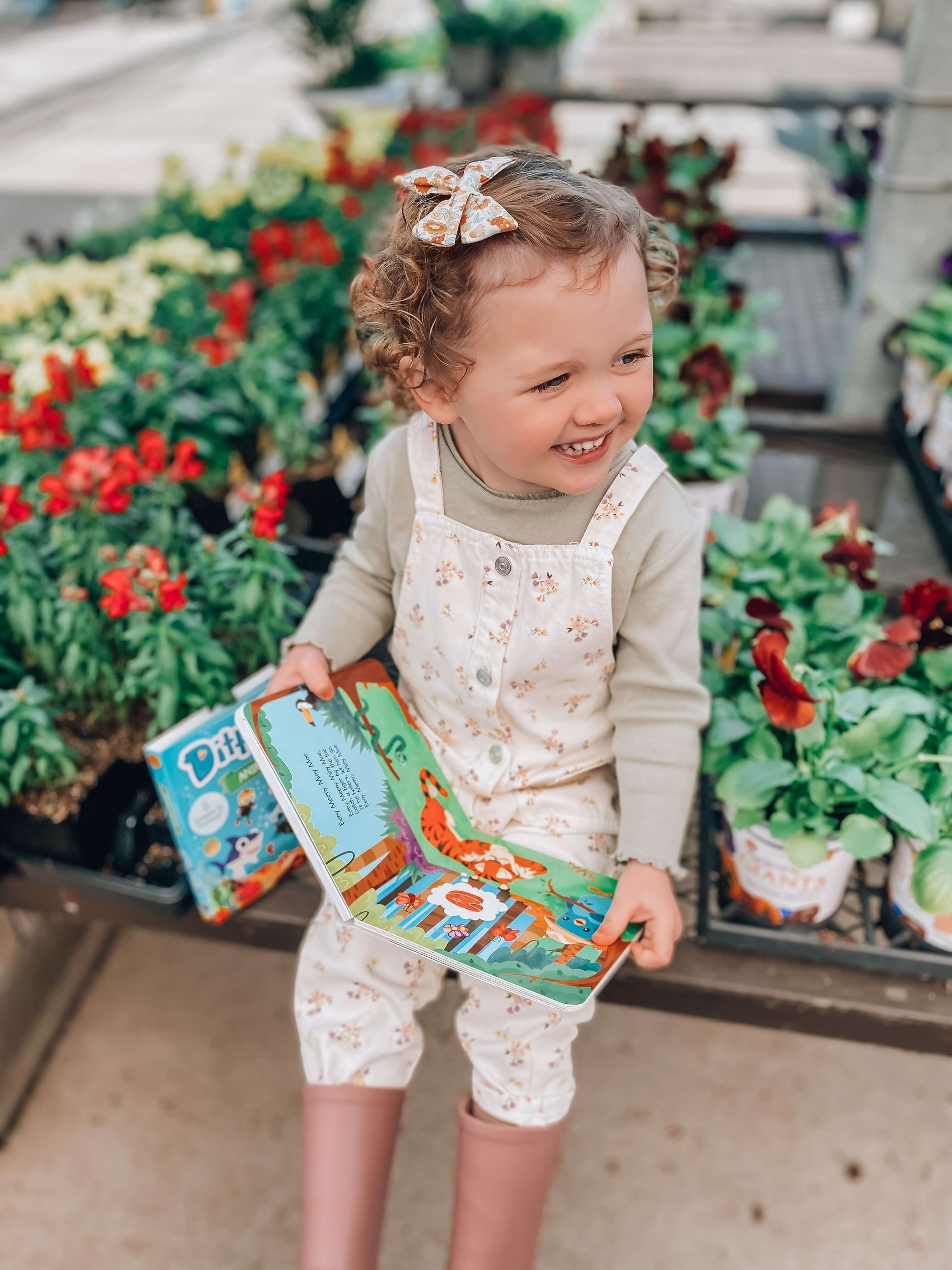 A smiling toddler with curly hair, wearing a bow, light overalls, and pink boots, sits among colorful potted flowers, holding an open Ditty Bird "Funny Songs" music book.