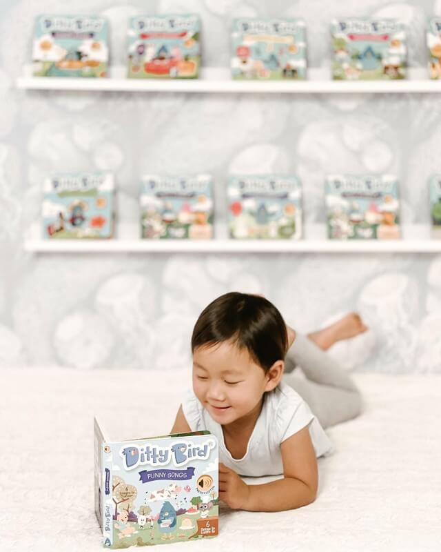 A young child relaxes on a bed, smiling while reading Ditty Bird’s Funny Songs toddler music book. More Ditty Bird titles are displayed on shelves in the background, adding to the bright and playful atmosphere.
