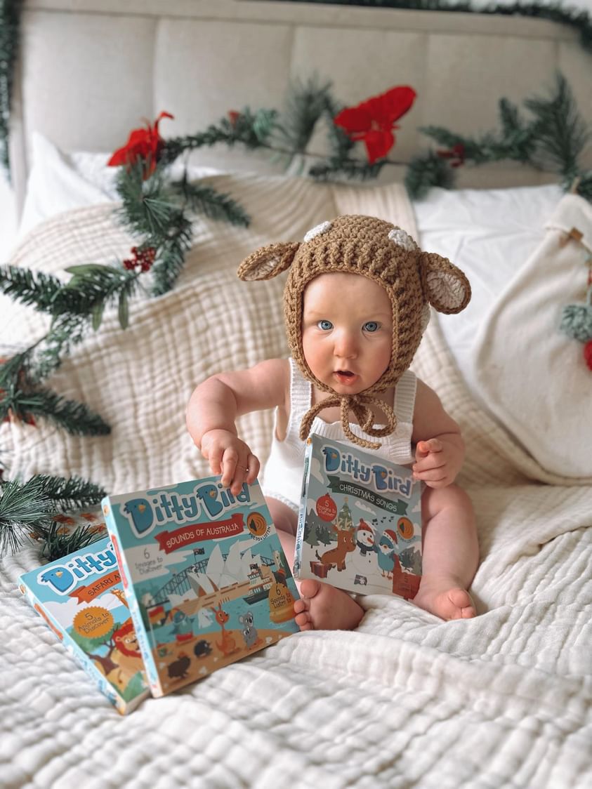 A baby in a knitted animal hat sits on a bed with festive garlands, holding the Ditty Bird "Sounds of Australia" book, surrounded by two more Ditty Bird books featuring wallabies and koalas.